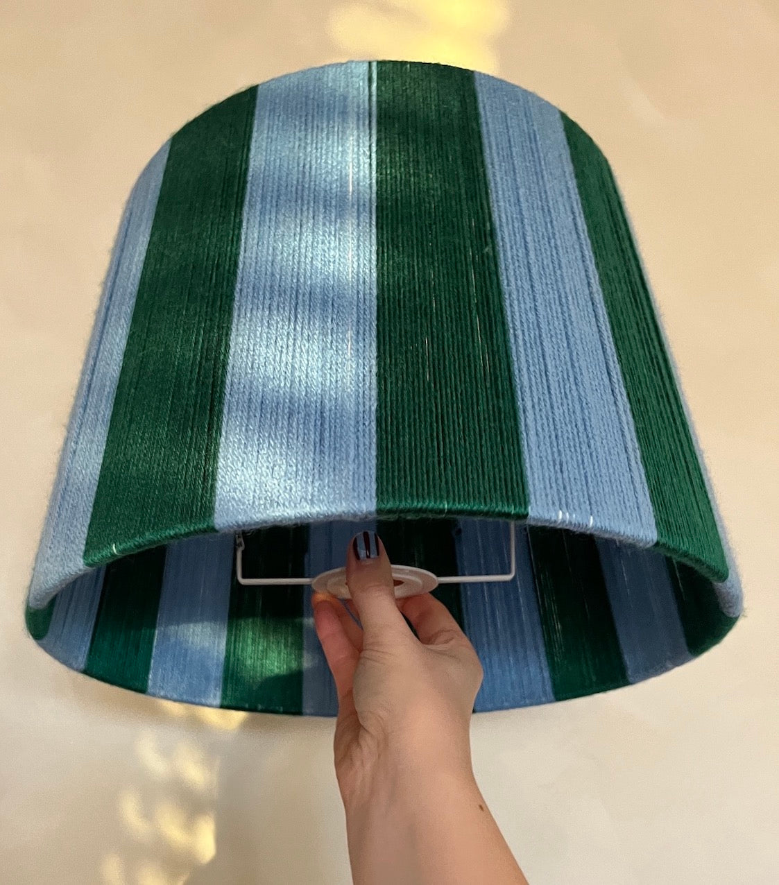 A hand is shown holding the bottom edge of a  Slice Shades handmade yarn drum lampshade with vertical stripes in forest green, light blue, and white, set against a warm beige background.