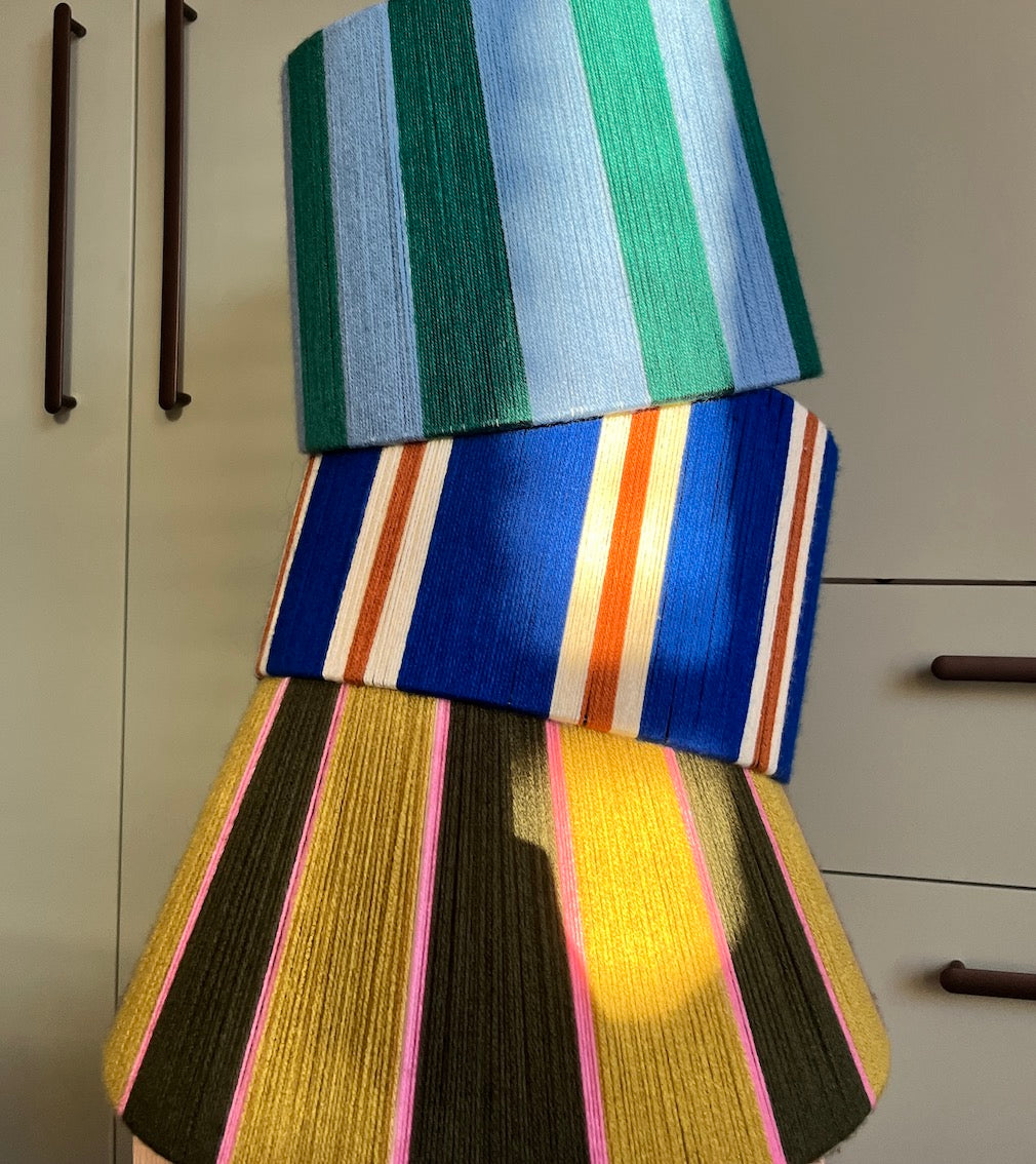 Three Slice Shades yarn ampshades stacked vertically, each featuring a distinct striped pattern in various colors including green, blue, yellow, pink, and orange, set against a neutral indoor background.