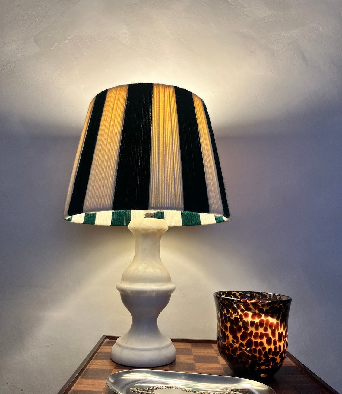 A Slice Shades table lamp with a white pedestal base and a string drum shade featuring vertical  green and white stripes, shown lit on a wooden side table next to a patterned candle.