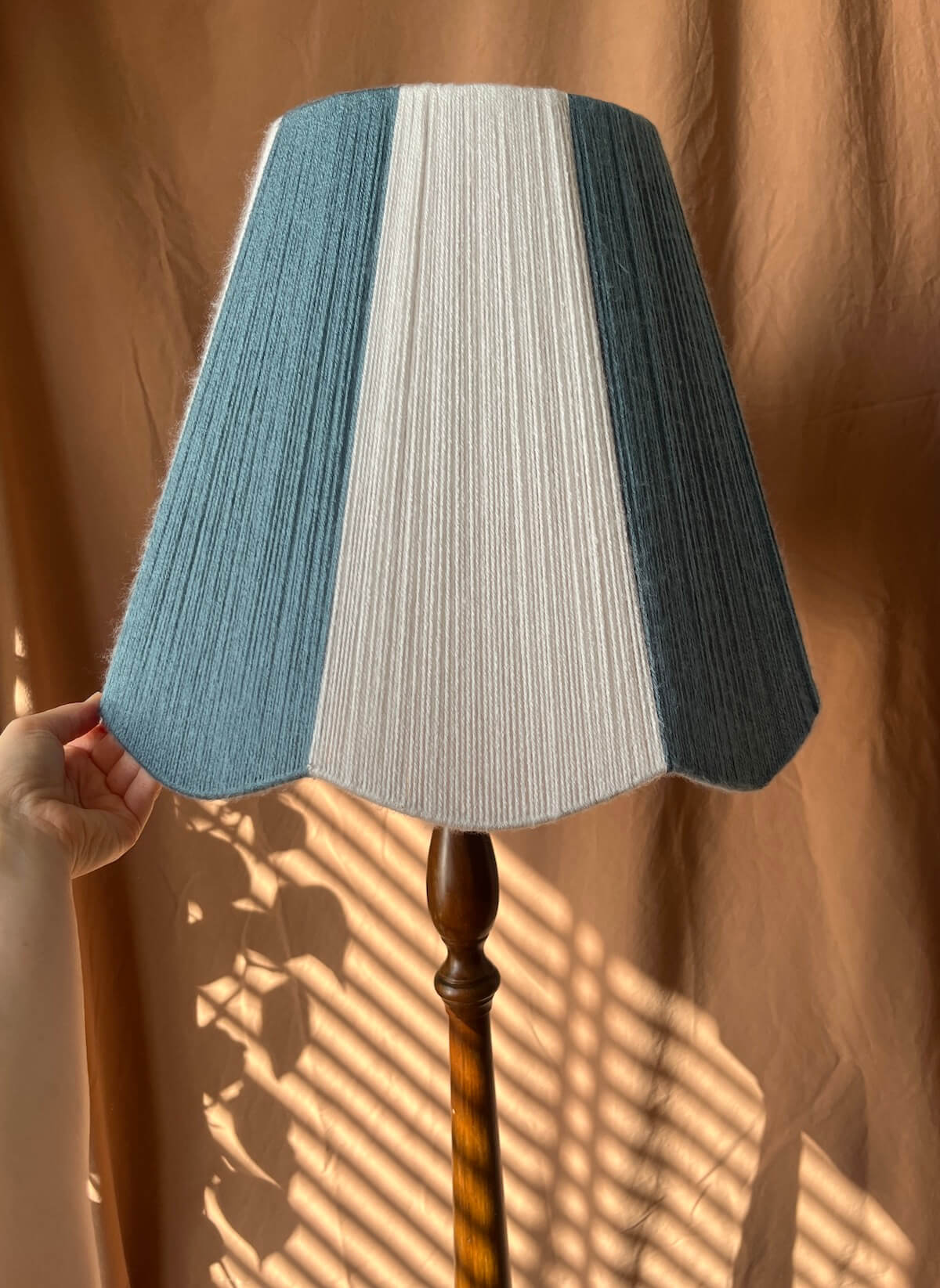 A Slice Shades scallop yarn lampshade with vertical stripes of off-white and slate blue, attached to a dark brown wooden floor lamp stand. A hand is holding the left edge of the shade, set against a draped brown curtain background with warm sunlight and leaf shadows.