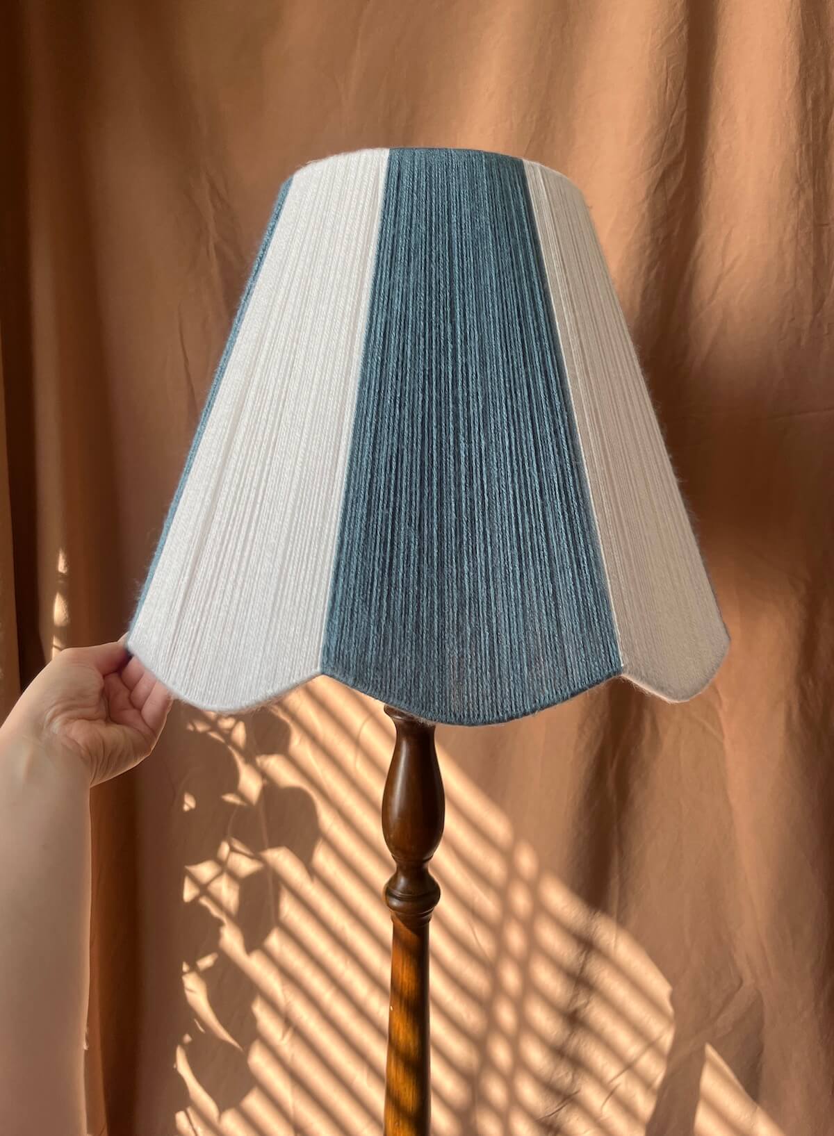 A Slice Shades scallop yarn lampshade with vertical stripes of off-white and slate blue, attached to a dark brown wooden floor lamp stand. A hand is holding the left edge of the shade, set against a draped brown curtain background with warm sunlight and leaf shadows.