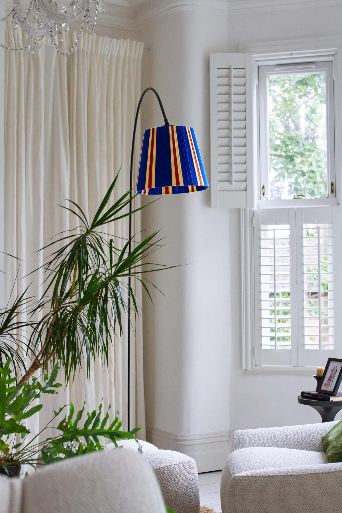A modern black floor lamp with a slice shades majorelle blue striped yarn lampshade, styled in a room with a cream curtain and green plants.