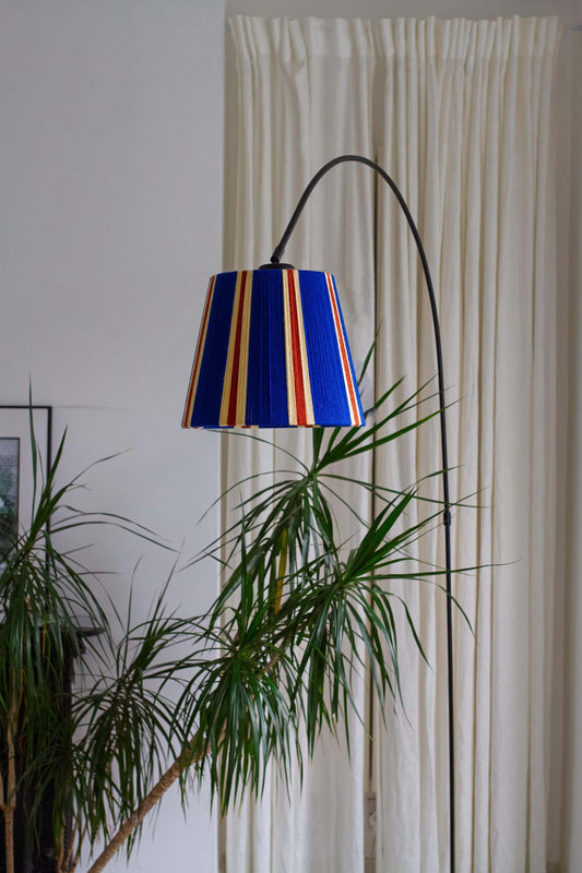 A modern black floor lamp with a slice shades majorelle blue striped yarn lampshade, styled in a room with a cream curtain and green plants.