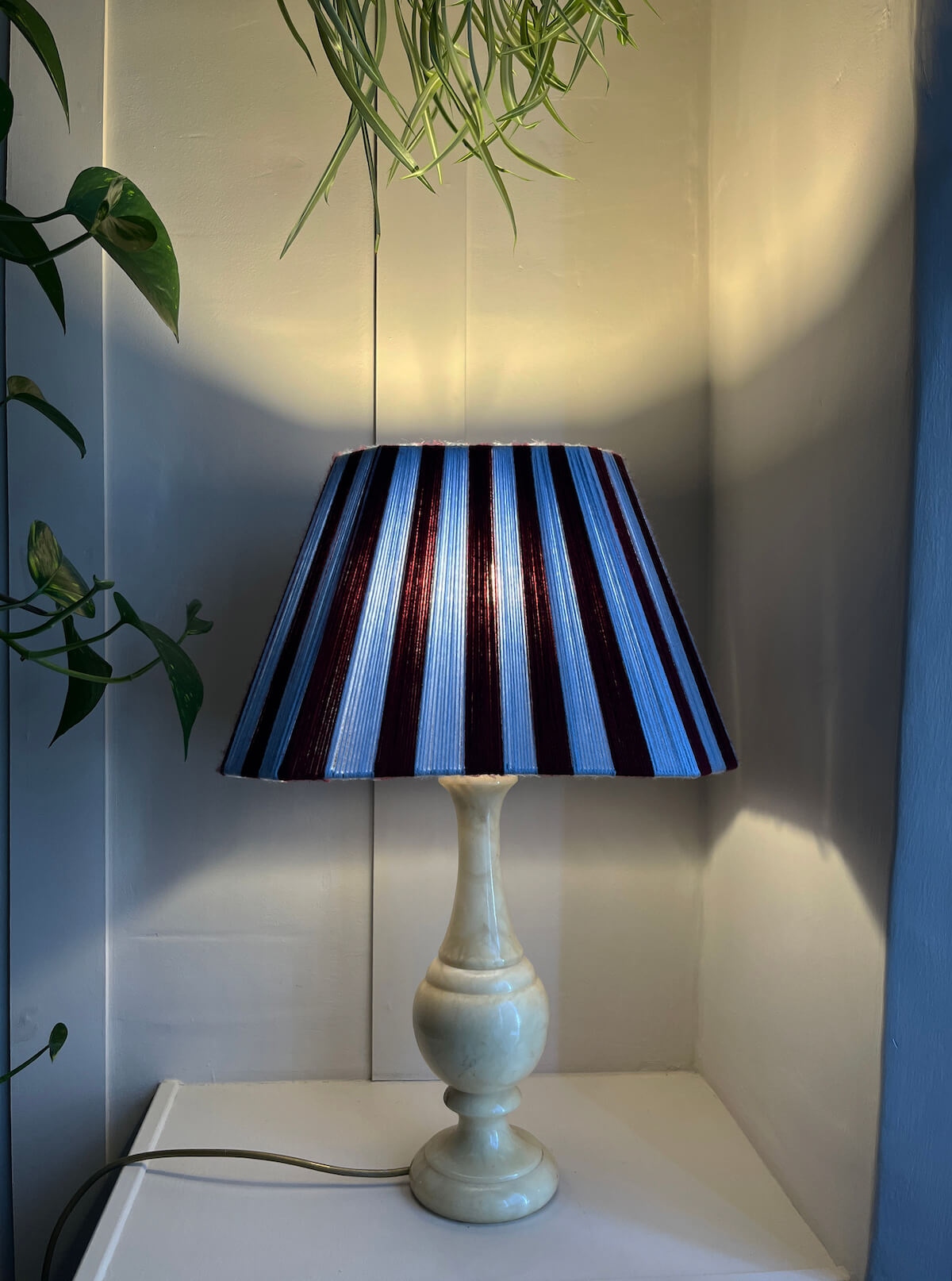 A table lamp with a Slice Shades yarn lampshade featuring vertical burgundy and sky blue stripes, mounted on a glossy pale yellow ceramic base. Styled in a corner roo and switched on and glowing.