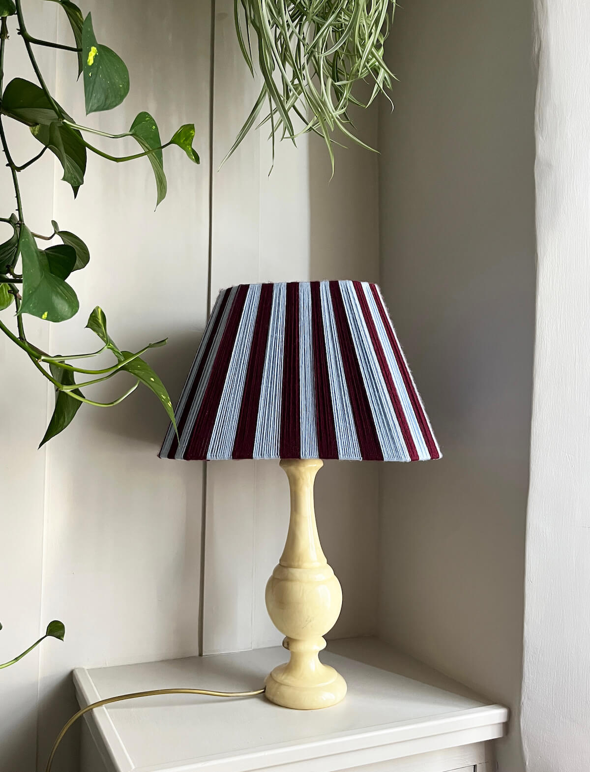 A table lamp with a Slice Shades yarn lampshade featuring vertical burgundy and sky blue stripes, styled in front of a panelled wall with plants.