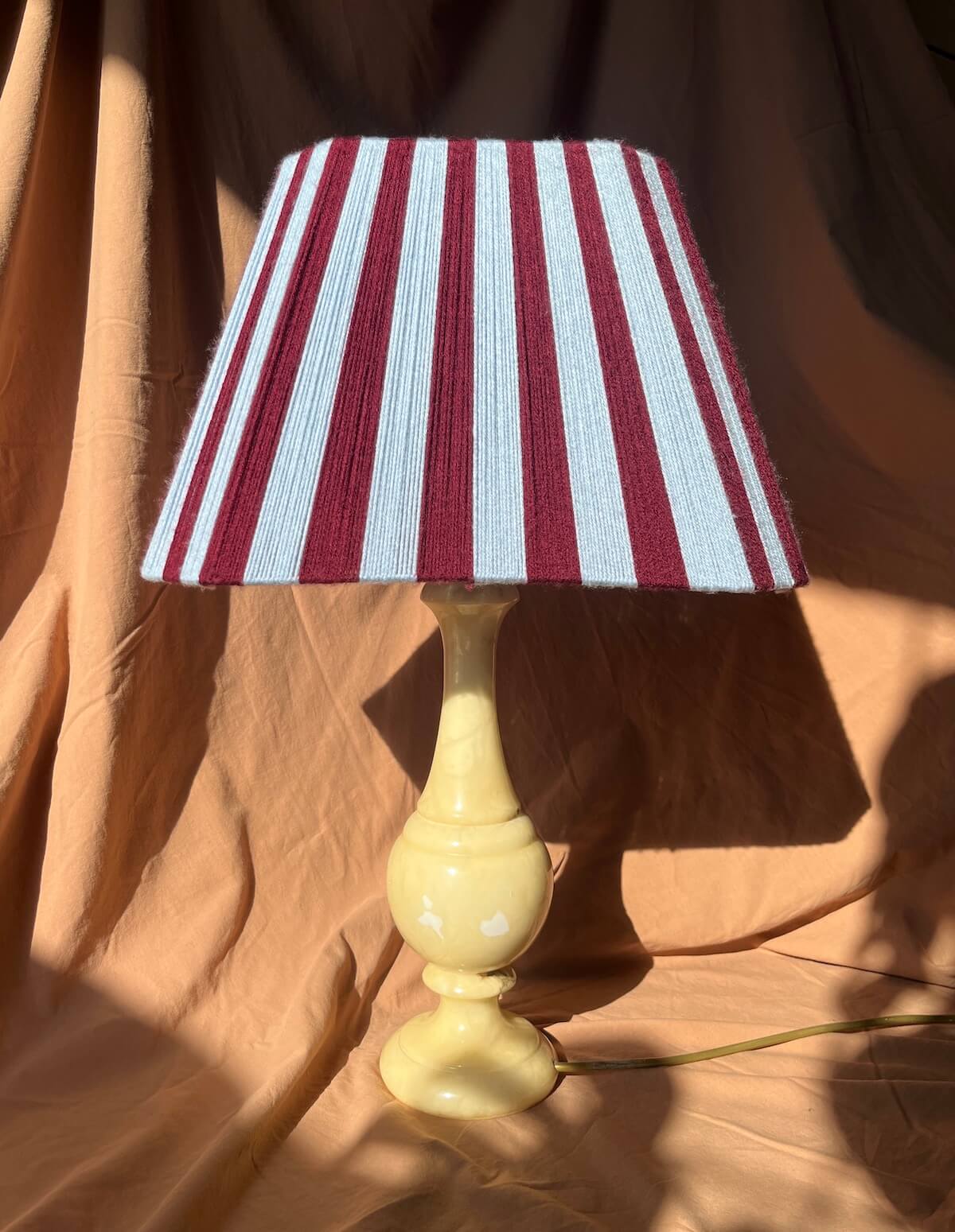 A table lamp with a Slice Shades yarn lampshade featuring vertical burgundy and sky blue stripes, mounted on a glossy pale yellow ceramic base, set against draped tan fabric.