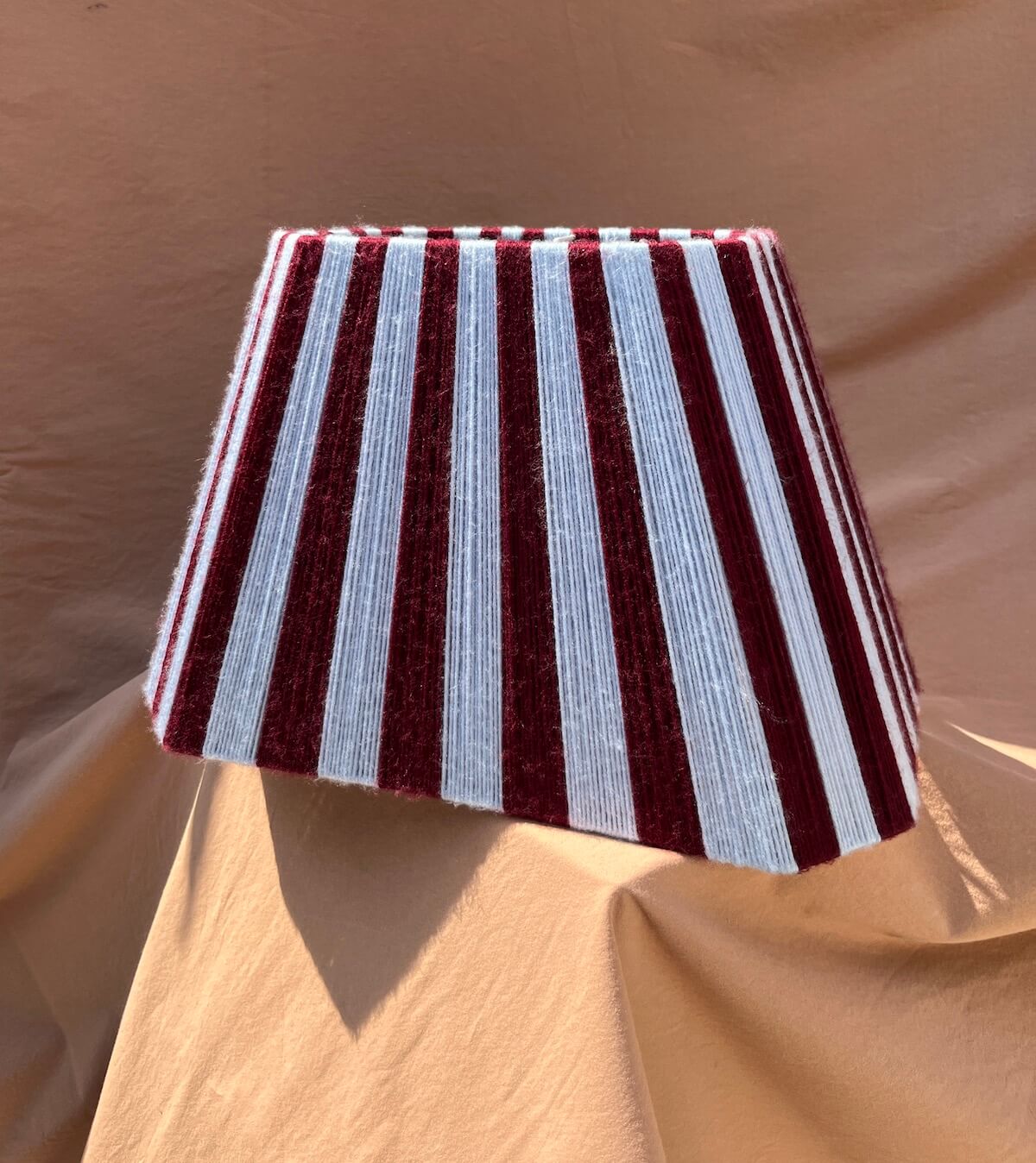 A Slice Shades yarn lampshade lampshade with alternating vertical stripes of deep burgundy and light blue, displayed against a draped tan fabric backdrop.