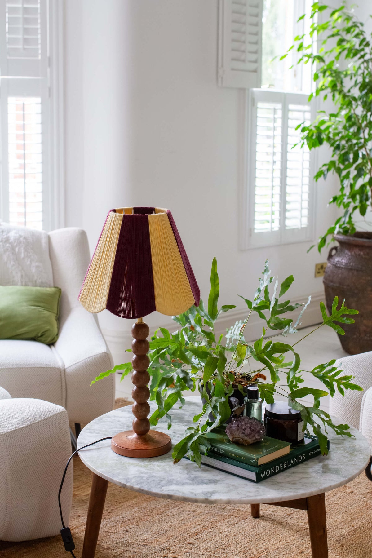 A mid-century style table lamp with  slice shades yarn scallop shade in maroon and yellow, a wooden bead spindle base, and a round wooden footed base, styled on a marble-top coffee table in a bright living room.
