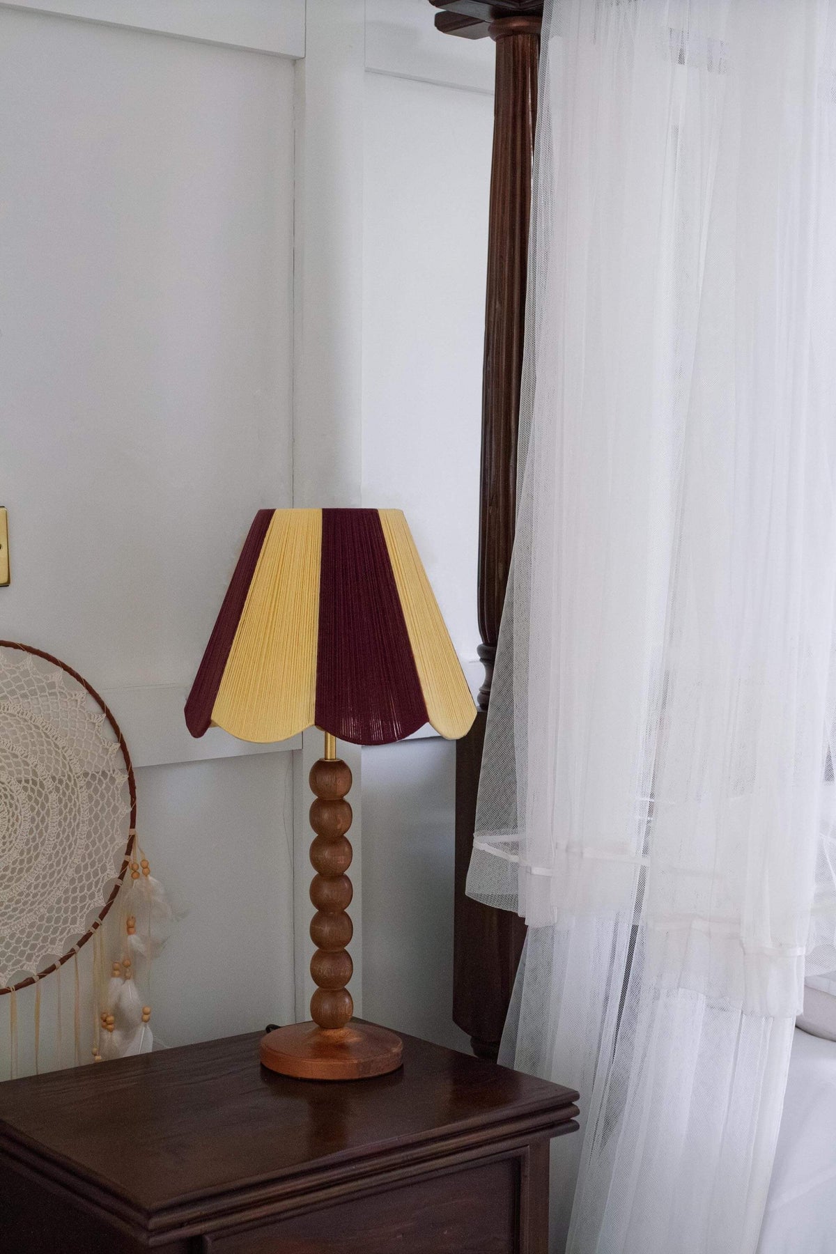 A table lamp with a slice shades yarn scallop shade in alternating dark burgundy and buttermilk yellow panels, supported by a round wooden pedestal base, set on a dark wood nightstand next to a window with white sheer curtains.