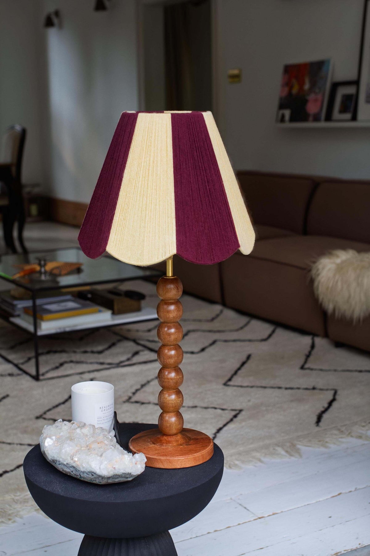 A slice shades yarn scallop lampshade with vertical stripes of buttermilk and deep burgundy, supported by a round wooden bead base on a dark pedestal table in a living room setting.