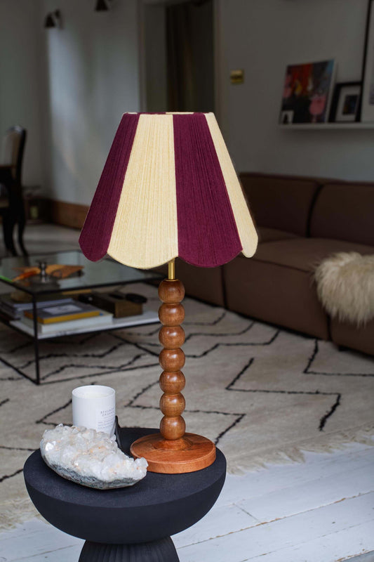 A slice shades yarn scallop lampshade with vertical stripes of buttermilk and deep burgundy, supported by a round wooden bead base on a dark pedestal table in a living room setting.