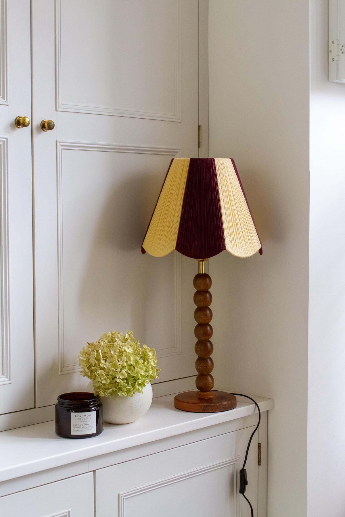 A table lamp with a slice shades yarn scallop shade in deep burgundy and pale yellow, mounted on a round wooden base, styled on a white sideboard next to a small potted plant and candle.