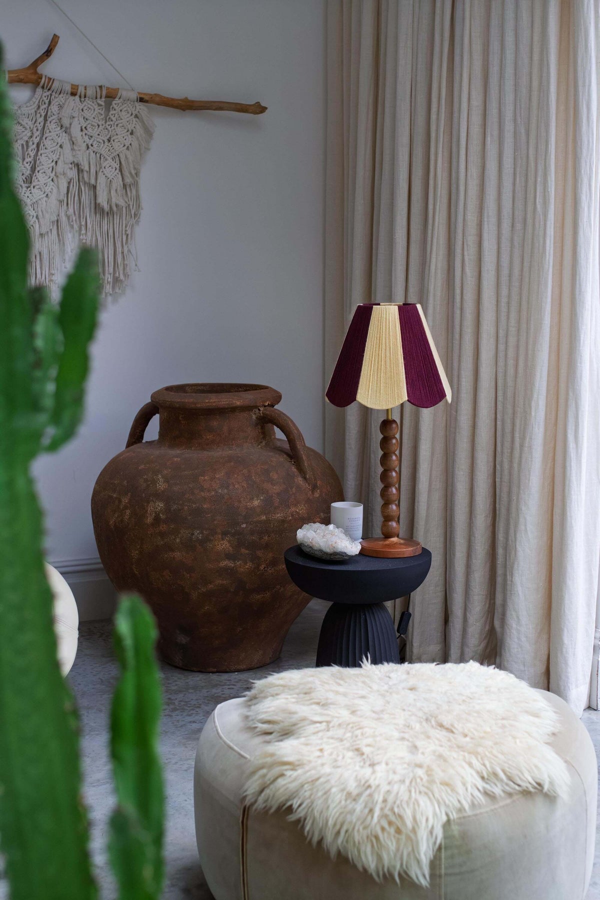 A styled interior corner with a large rustic terracotta vase, a small black side table holding a striped slice shades lamp, a white fluffy ottoman, cream curtains, and a macrame wall hanging.