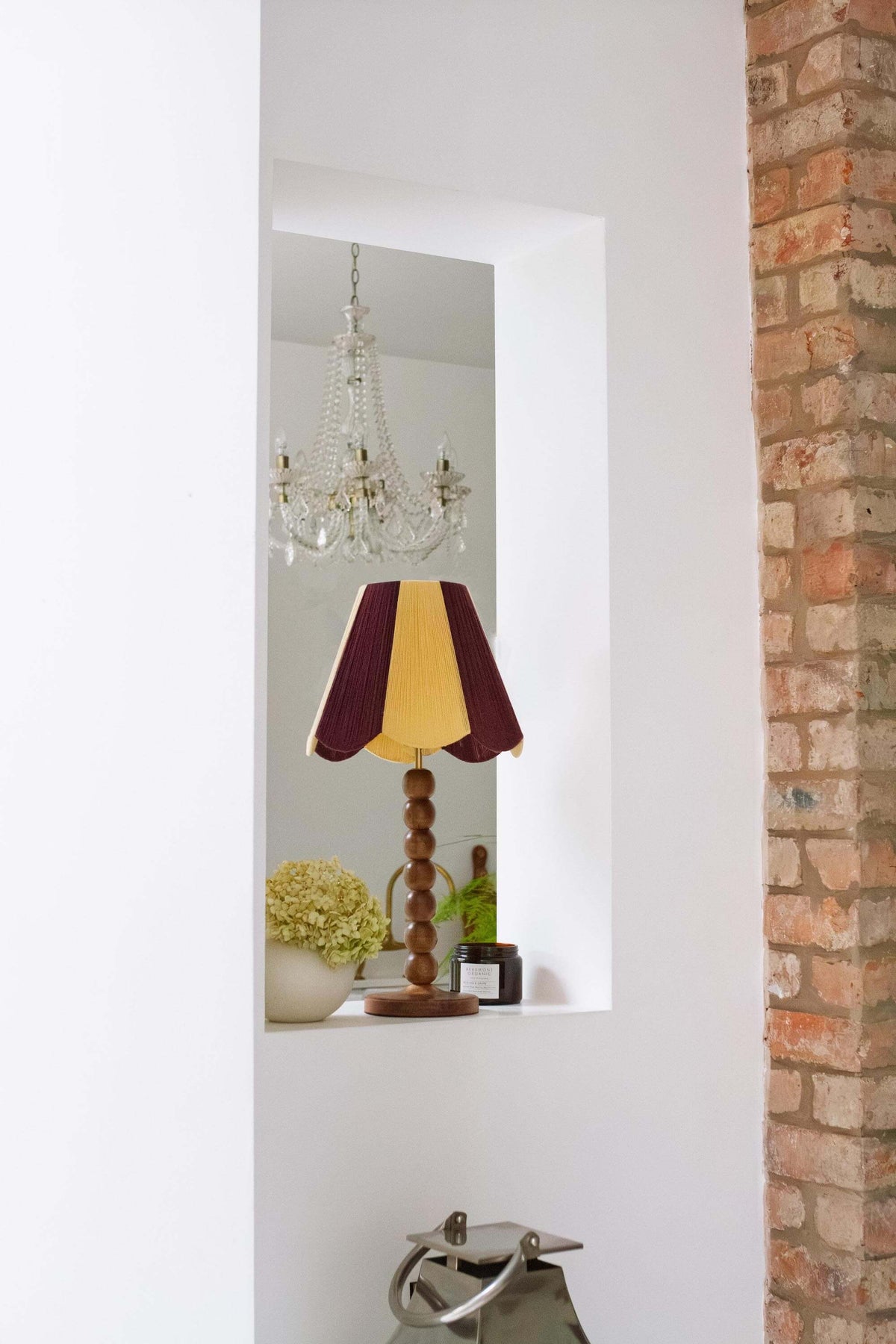 A slice shades yarn scallop shade with a maroon and yellow striped shade and a turned wooden base, displayed on a shelf in a white wall niche next to a brick column.