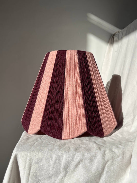 A Slice Shades handwoven yarn lampshade with vertical stripes in dusty pink  and deep burgundy, set against a draped white fabric background.