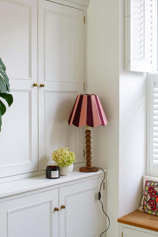 A pink and burgundy slice shades handwoven yarn lampshade table lamp with a wooden bead stem base, styled on a white wooden credenza in a bright room.