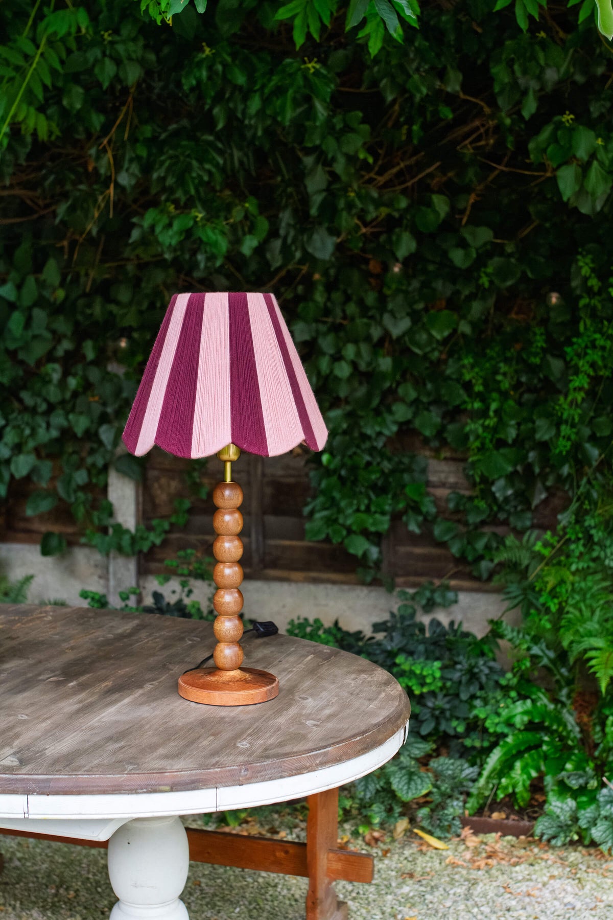 A table lamp with a pink and purple striped slice shades handwoven yarn lampshade and a round wooden pedestal base, set on a rustic outdoor table with a lush green garden background.