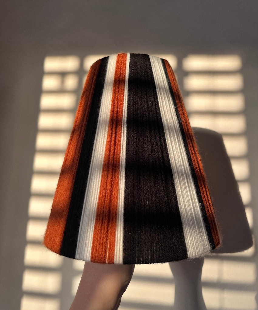A Slice Shades handwoven yarn lampshade with vertical stripes of rust orange, black, and off-white, displayed against a background with soft window shadow patterns.