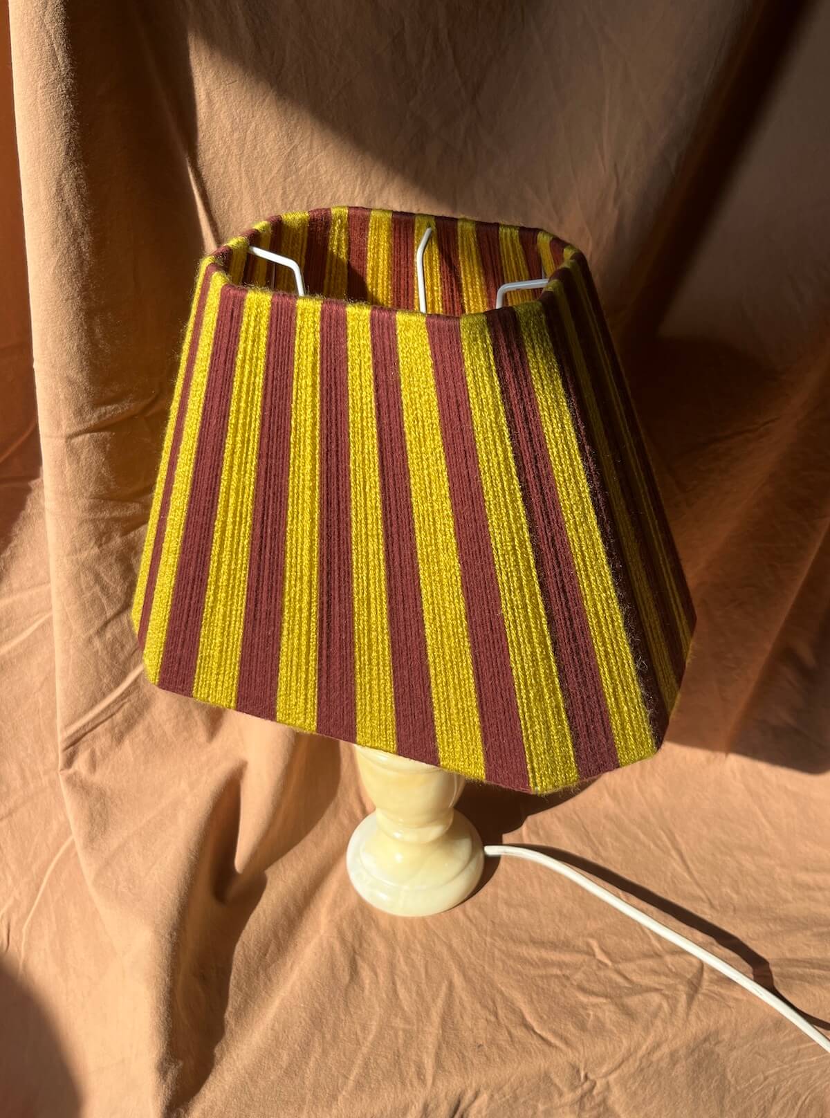 A table lamp with a Slice Shades yarn lampshade featuring vertical stripes of chartreuse and burgundy, mounted on a cream-coloured ceramic base. The lamp is photographed against a draped tan fabric backdrop with direct sunlight casting shadows.