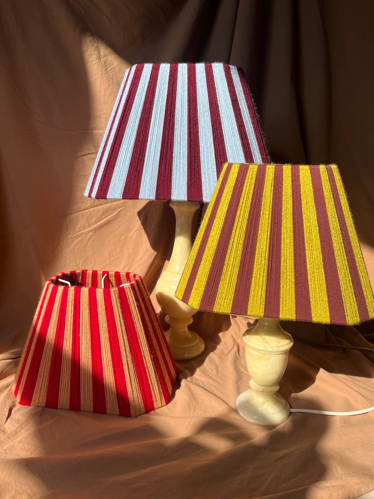 Three striped yarn Slice Shades lampshades and matching lamp bases displayed on draped tan fabric under warm directional sunlight. The shades and bases feature vertical stripes in red, cream, yellow, and dark red/burgundy.