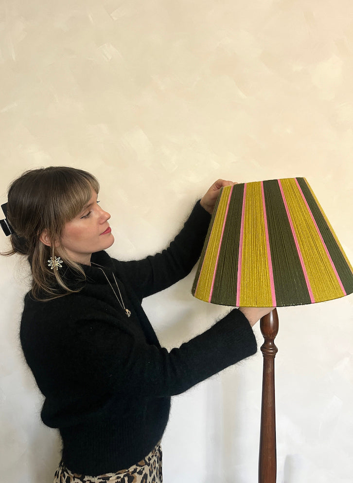 A woman in a black sweater adjusts a Slice Shades yarn floor lampshade with vertical stripes of olive green, mustard yellow, and pink, set against a textured off-white wall.