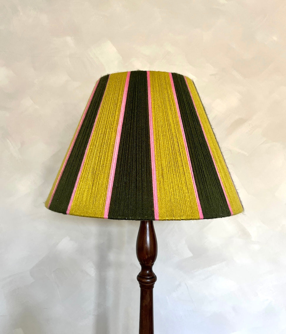 A Slice Shades yarn lampshade with vertical stripes of mustard yellow, dark olive green, and pink, mounted on a dark brown wooden stand against a textured off-white wall.