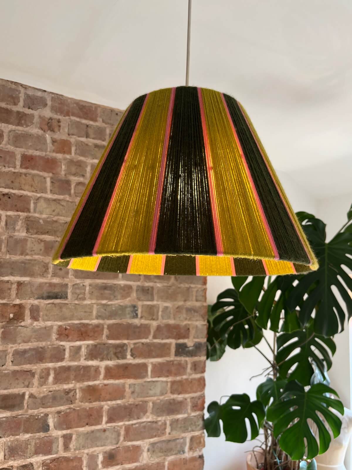 A glowing Slice Shades yarn tapered lampshade with vertical stripes of gold, dark brown, and pink hanging in a room with a brick wall, white ceiling, and a potted monstera plant. 