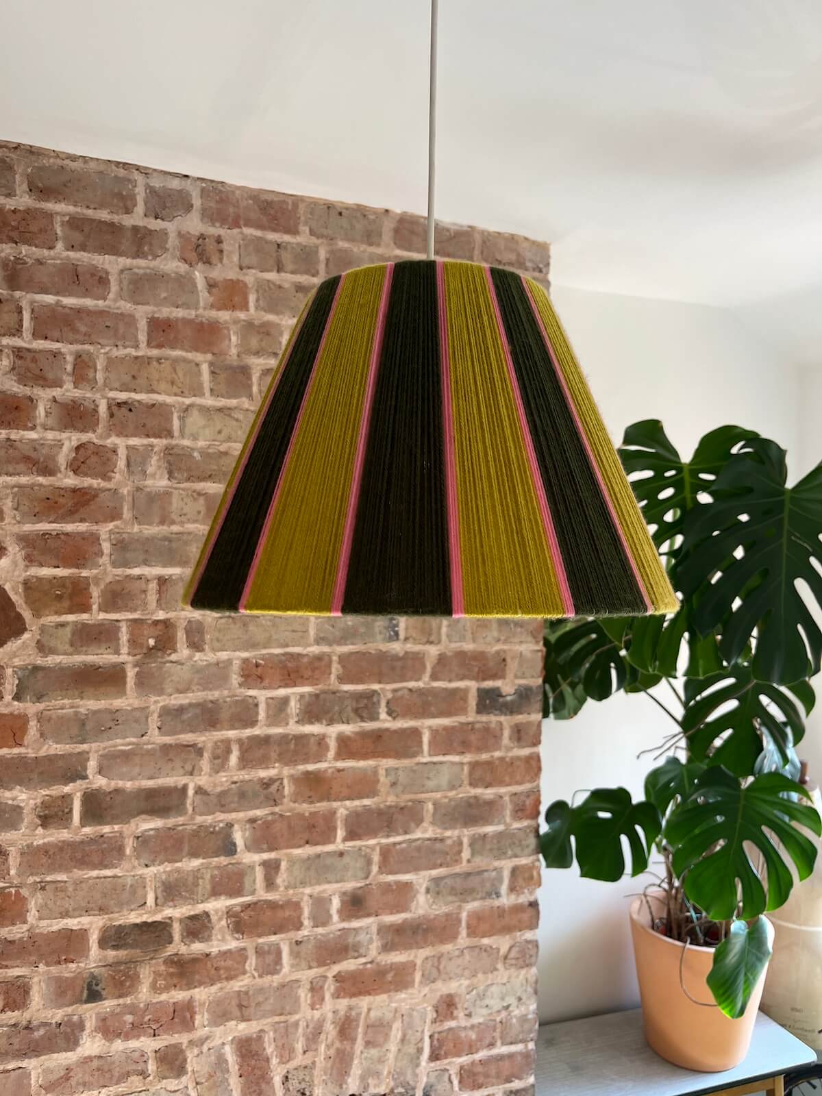 A Slice Shades yarn tapered lampshade with vertical stripes of gold, dark brown, and pink hanging in a room with a brick wall, white ceiling, and a potted monstera plant. 