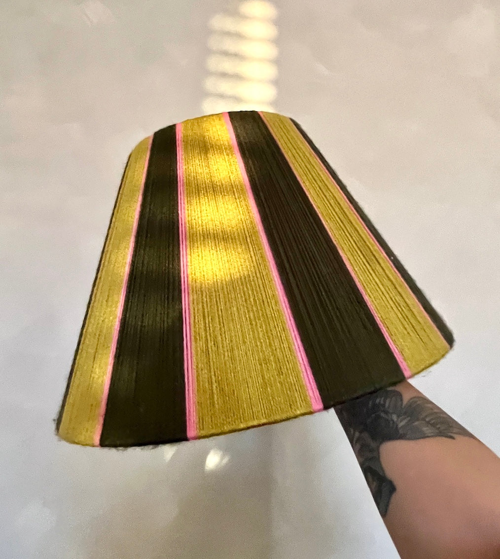 A Slice Shades yarn tapered lampshade with vertical stripes of gold, dark brown, and pink, held up by a tattooed arm against a light gray background.