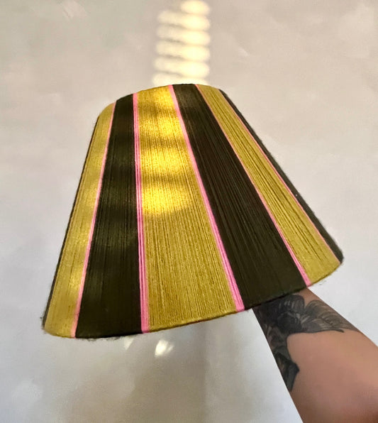 A Slice Shades yarn tapered lampshade with vertical stripes of gold, dark brown, and pink, held up by a tattooed arm against a light gray background.