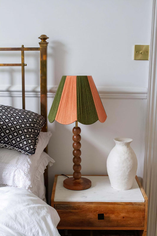 A mid-century style table lamp with a slice shades yarn lampshade in green and peach, set on a wooden bedside table next to a white ceramic vase in a bedroom setting.