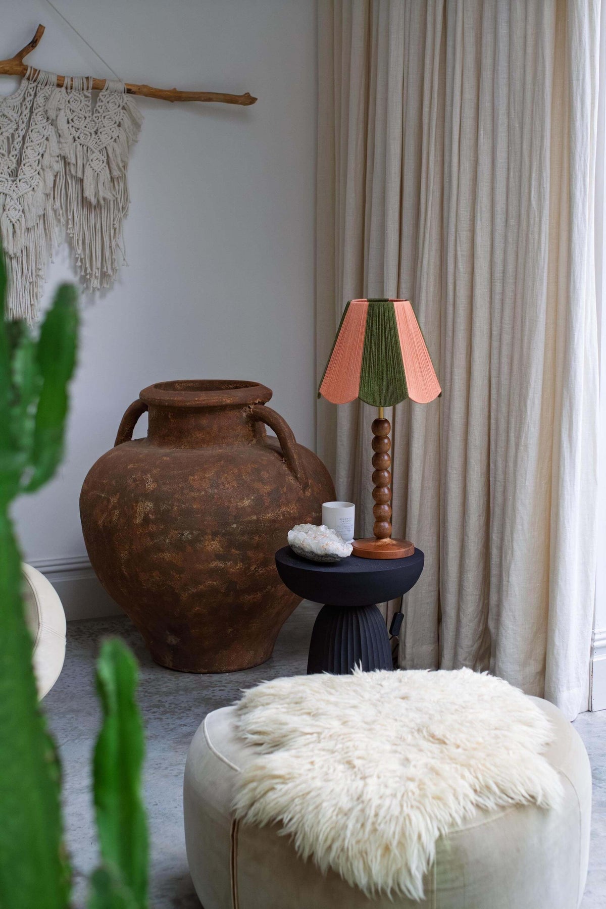 A styled interior corner featuring a large rustic brown clay urn, a black side table with a slice shades yarn lampshade in green and peach, a cream pouf with a white faux-fur throw, and beige curtains in the background.