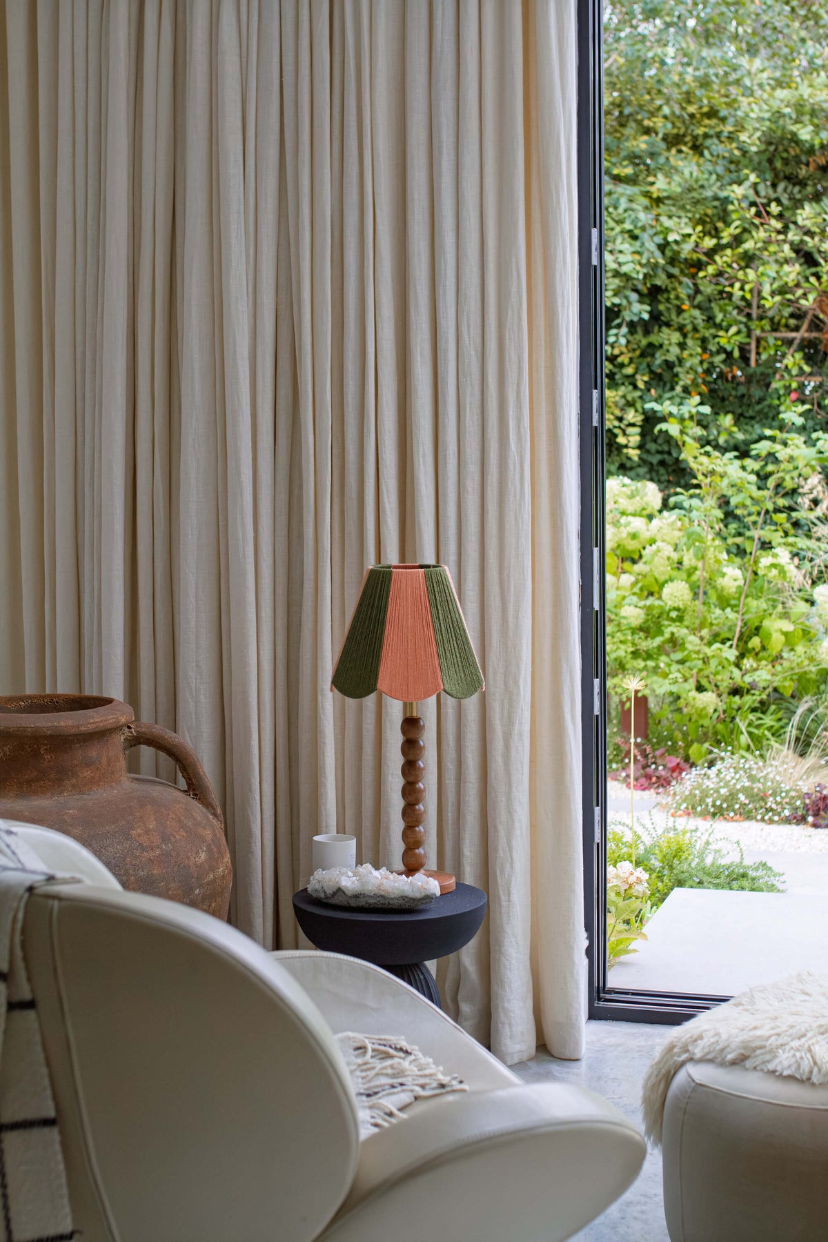 Beige pleated curtains hanging next to a large glass door that opens onto a lush green garden. In the foreground, a cream-colored armchair and a small side table with a slice shades yarn lampshade in green and peach are visible.