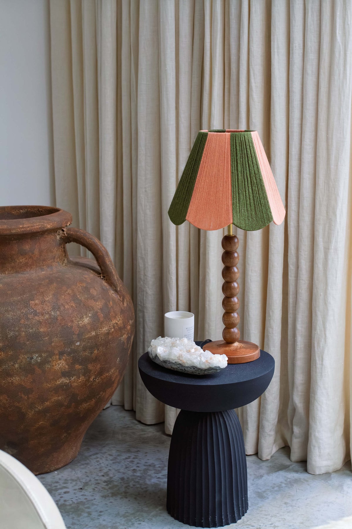 A decorative table lamp with a pslice shades yarn lampshade in green and peach, set on a black side table next to a large rustic brown vase against a backdrop of beige curtains.