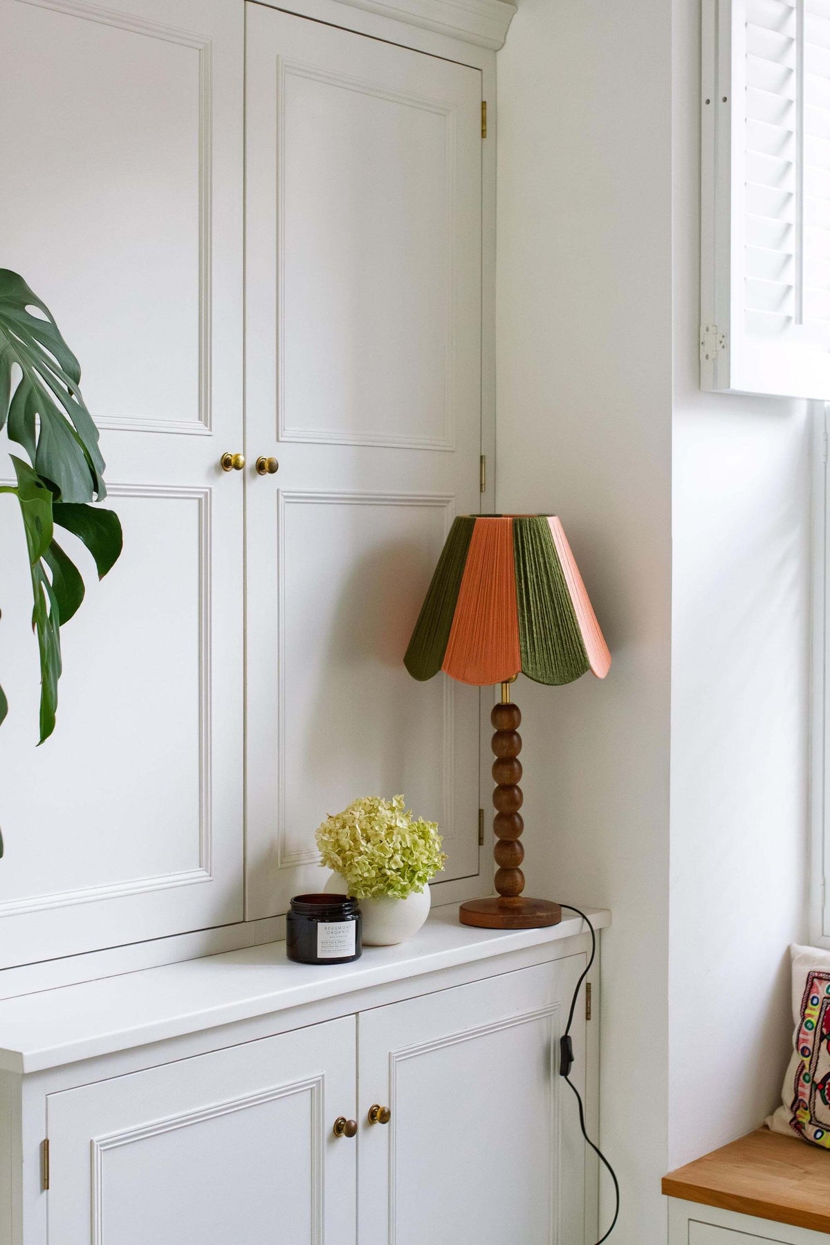 A decorative table lamp with a slice shades yarn lampshade in green and peach and a round wooden base, styled on a white cabinet in a bright room.