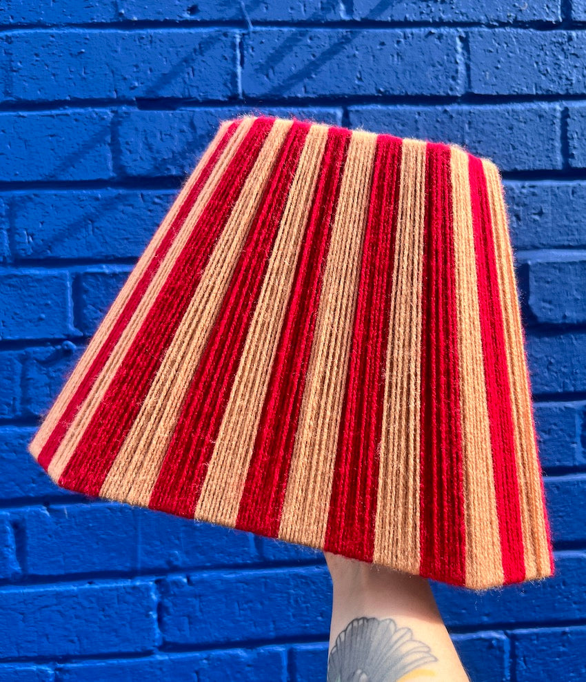 A handwoven string lampshade with vertical stripes of red and peach yarn, held up against a bright blue painted brick wall.