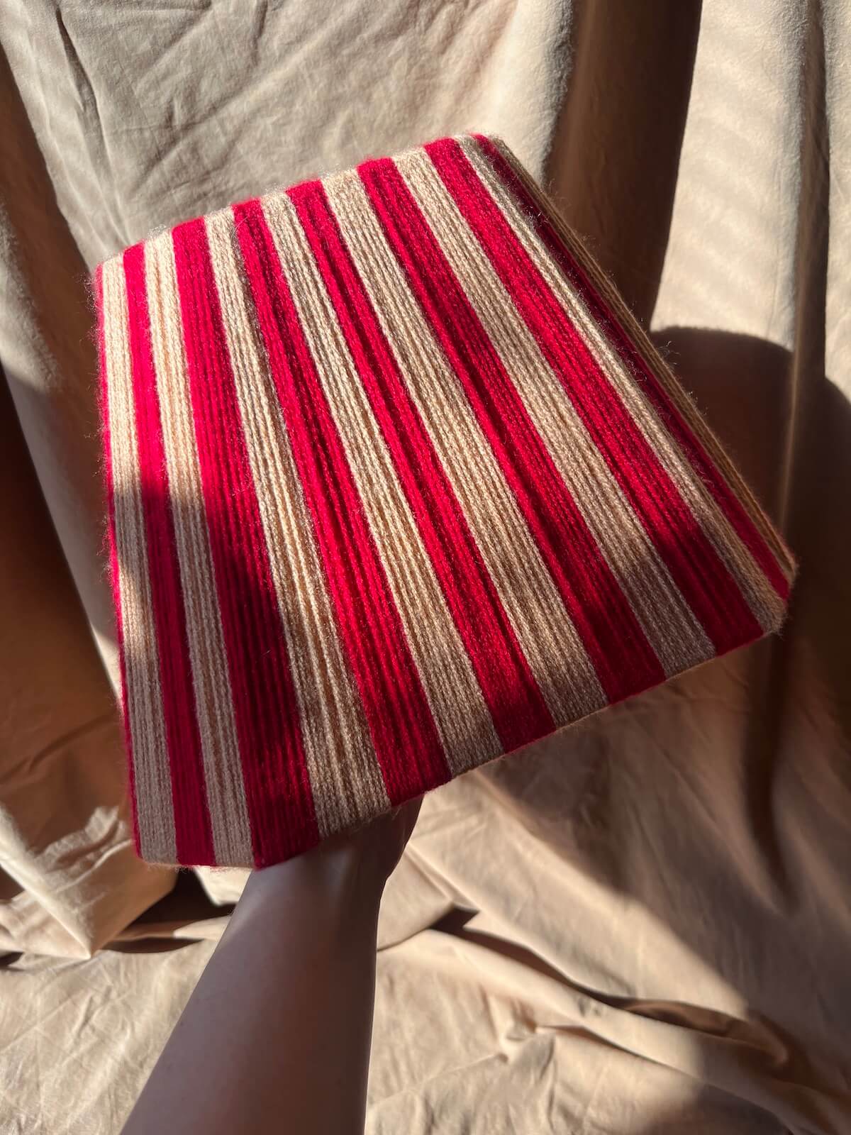 An octagonal string lampshade in red and peach held by a hand against a backdrop of draped beige fabric.