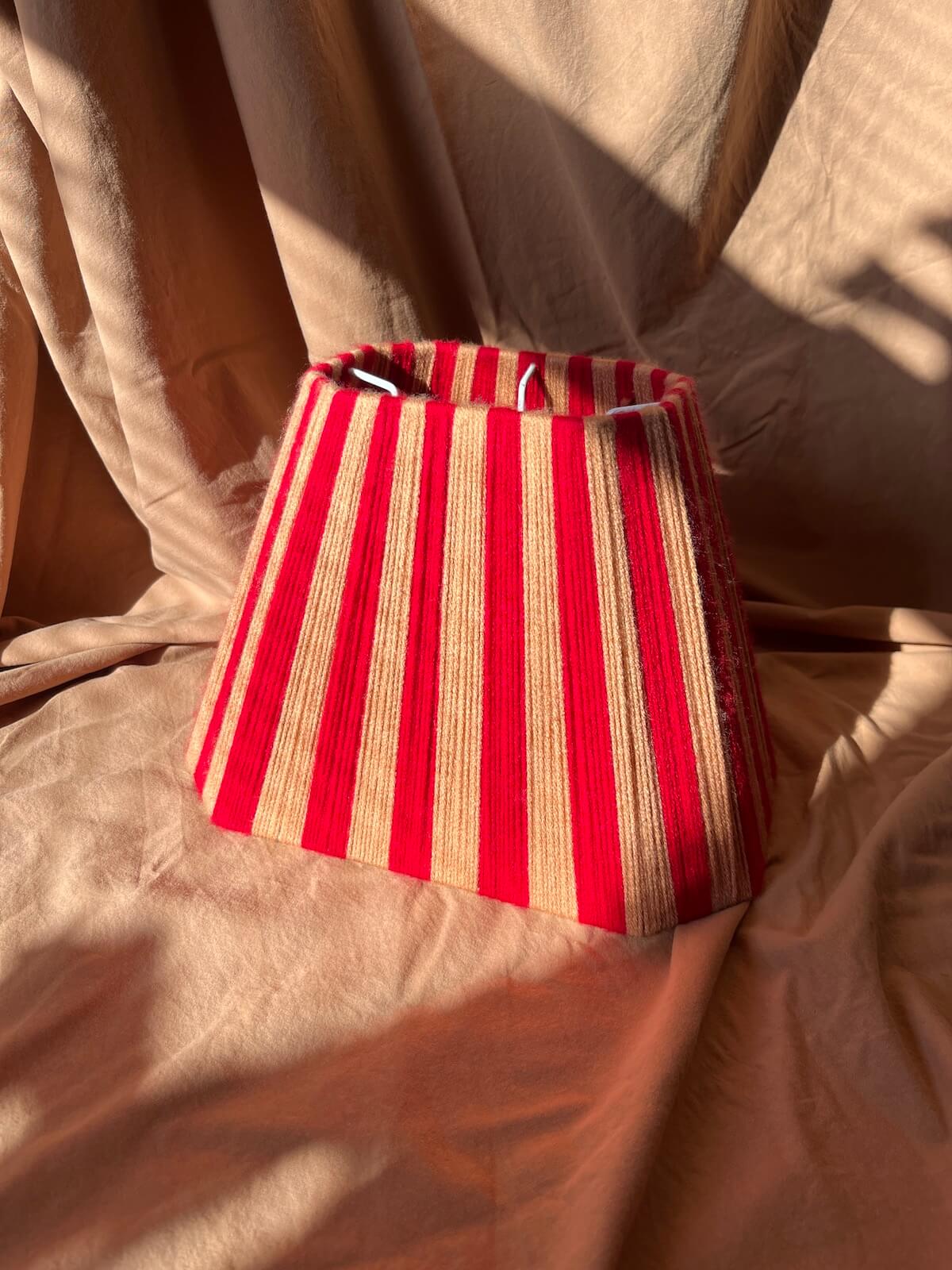 A fabric lampshade with vertical red and beige stripes, photographed on draped beige fabric under warm, direct sunlight with soft shadows.