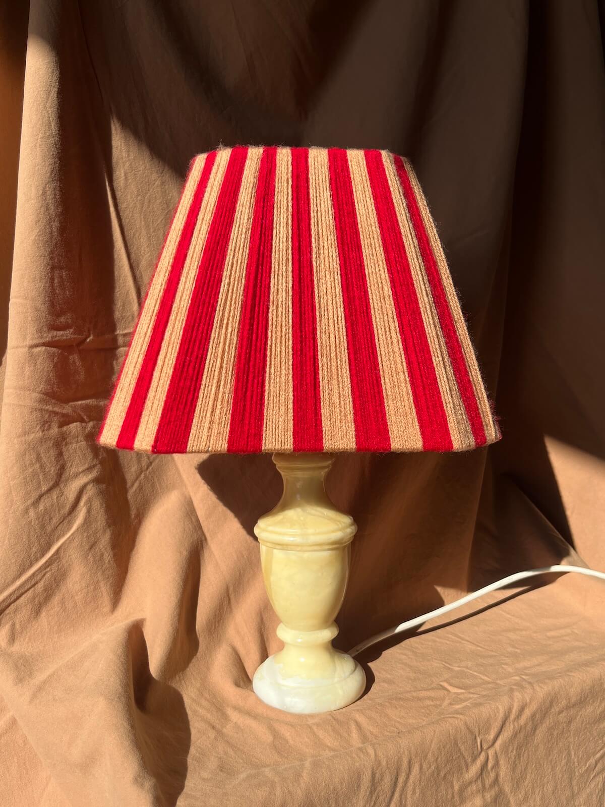 A table lamp with a striped shade featuring vertical red and peach stripes, mounted on a cream-colored stone or ceramic pedestal base, set against a draped brown fabric backdrop.