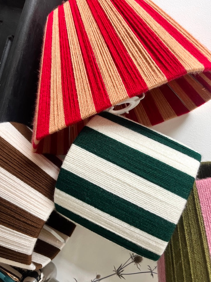 A close-up of striped string lampshades draped together, including a red and beige striped piece, a dark green and white striped piece, and other partial colourways at the edges.
