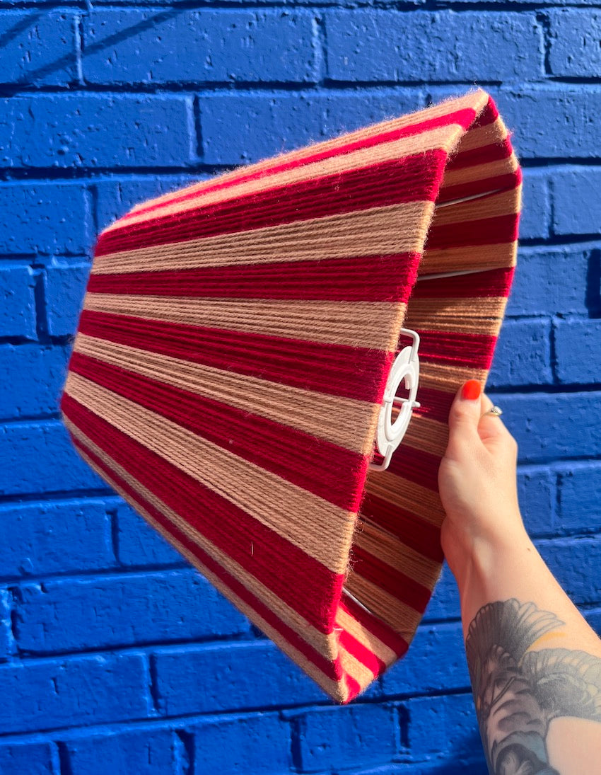 A hand holding a striped red and peach wool lampshade against a bright blue brick wall background.