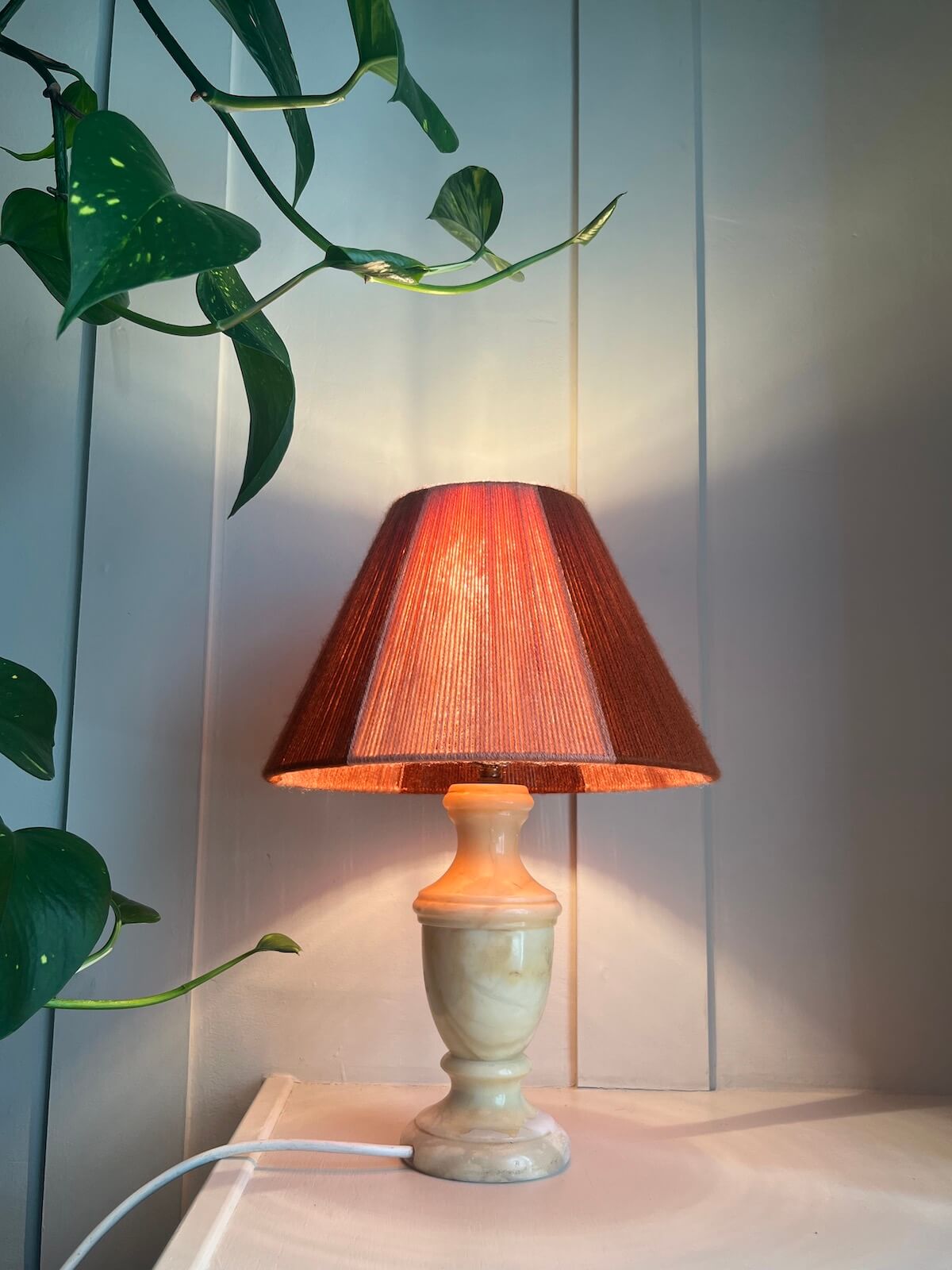 A Slice Shades table lamp with a pleated reddish-brown fabric shade and a cream-coloured stone base, illuminated and placed on a light surface against a panelled wall with green plant leaves entering from the top left.