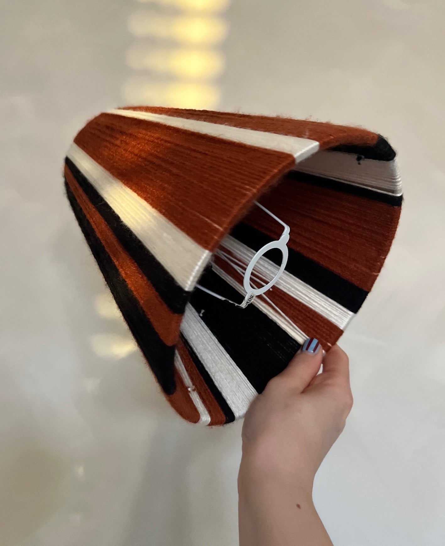 A Slice Shades handwoven yarn lampshade in multi width colours of black, rust and ecru held up in front of a beige background.