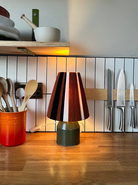 A modern table lamp with a Slice Shades yarn lampshade made of alternating vertical stripes of dark brown, black, white, and reddish-brown, sitting on a wooden kitchen counter.