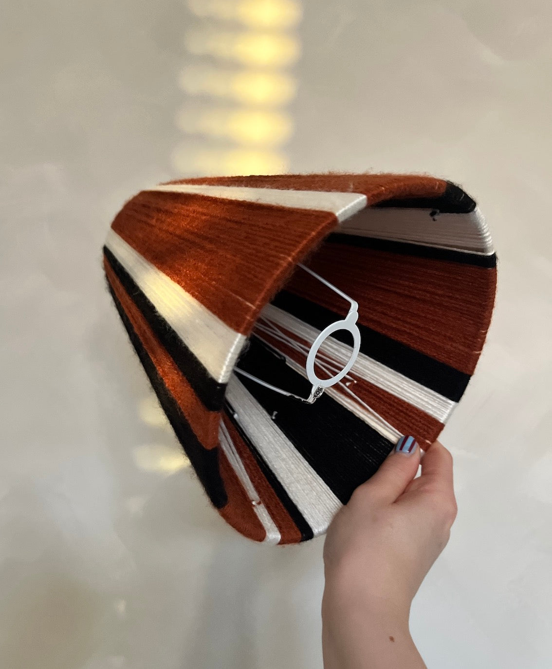 A hand holding a Slice Shades striped yarn lampshade with brown, white, and black stripes, set against a light-coloured background with soft window light reflections.