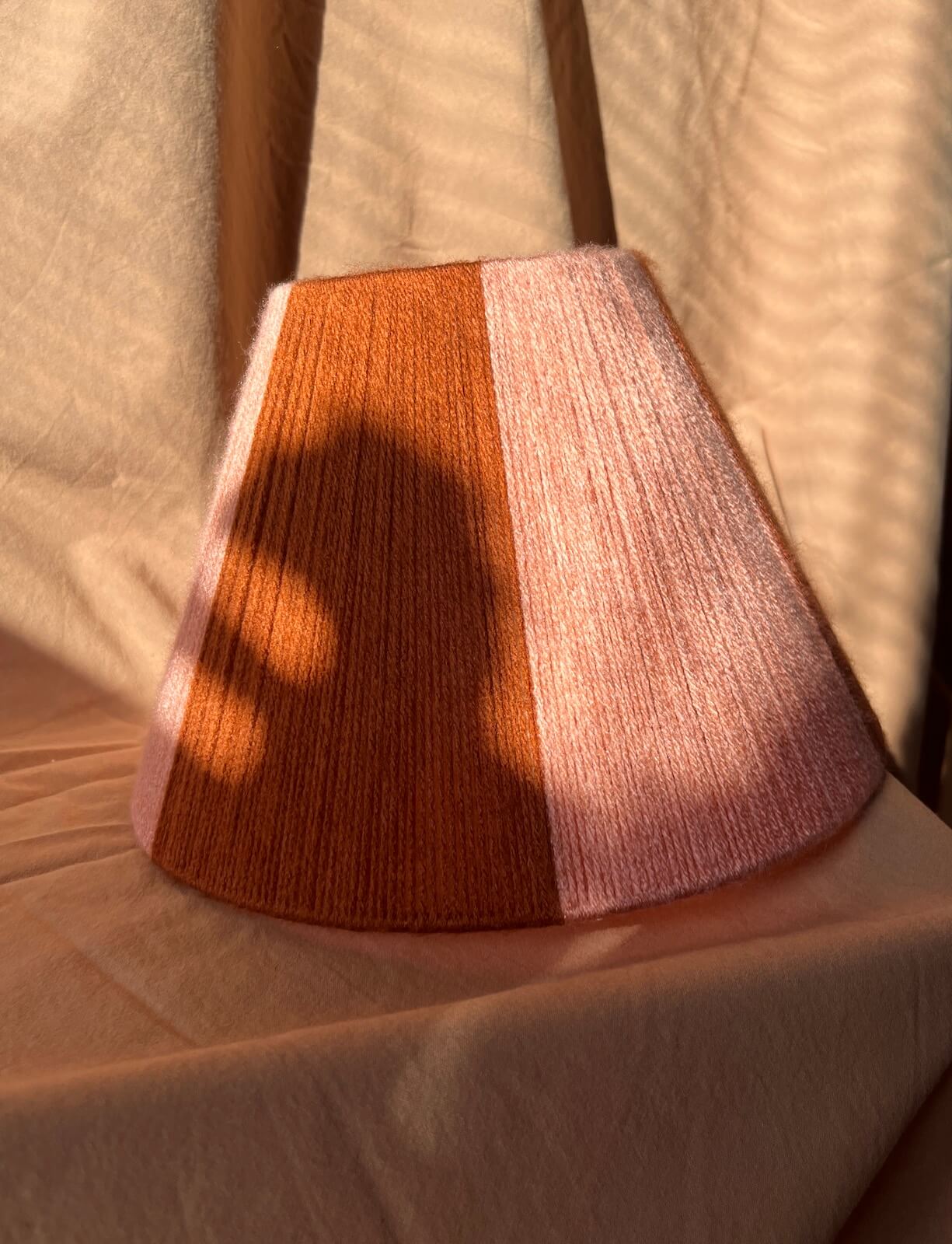 A string yarn lampshade with chunky vertical stripes in burnt orange and light pink, resting on draped beige fabric under warm, directional sunlight that casts a shadow across the lampshade.