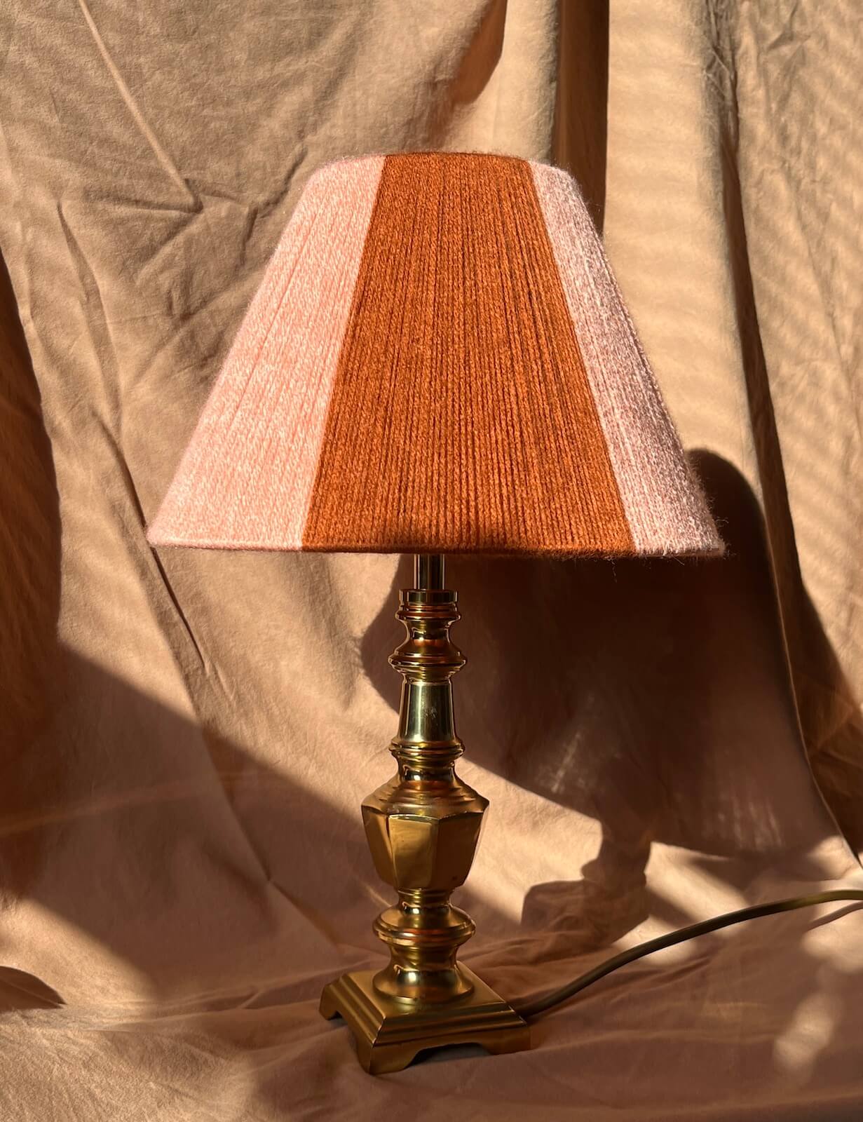 A vintage-style table lamp with a handowoven string shade in vertical stripes of rust orange and pale pink, mounted on a polished brass base and set against draped beige fabric.