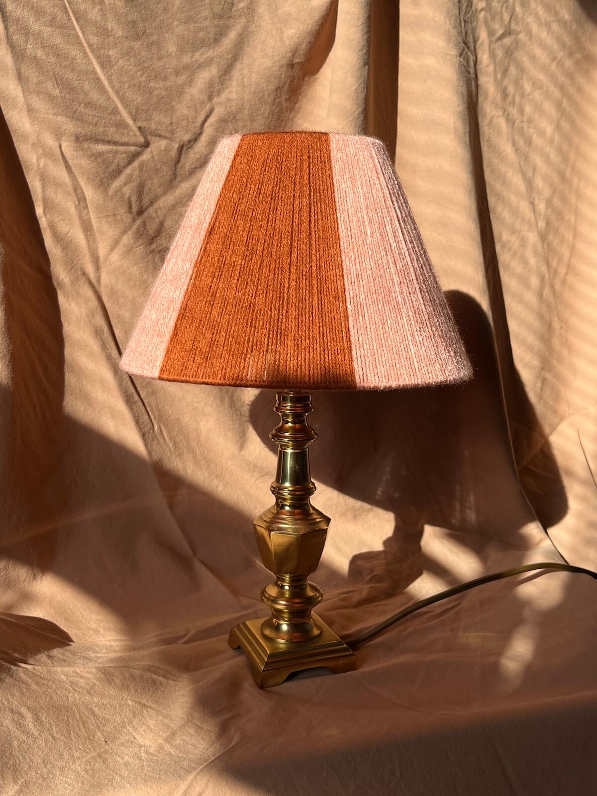 A table lamp with a stripey string shade in vertical stripes of rust orange and light pink, mounted on an ornate brass-colored metal base, set against a draped beige fabric backdrop.