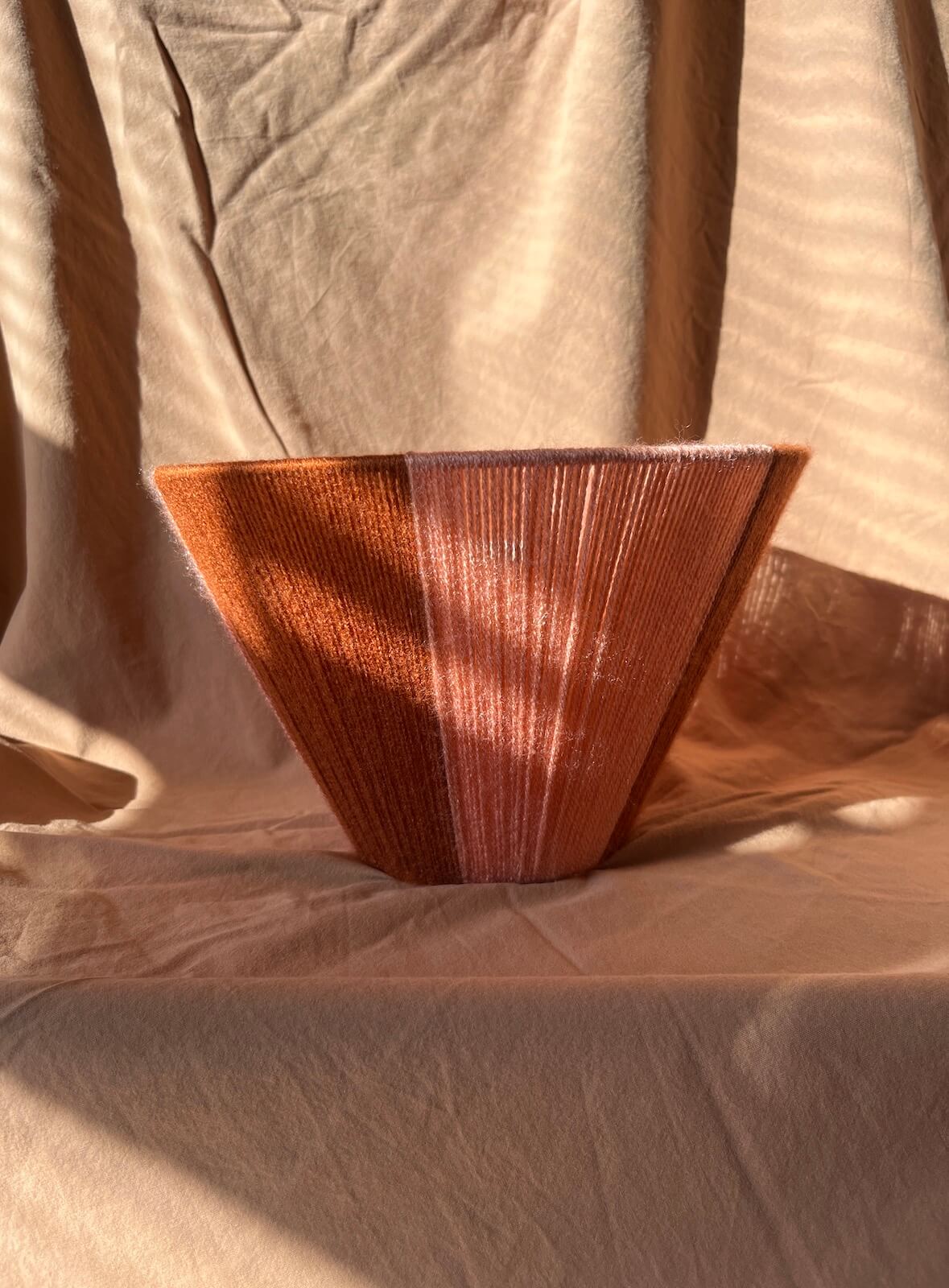 A handmade woven lampshade made of string in vertical pink and rust stripes upside down.