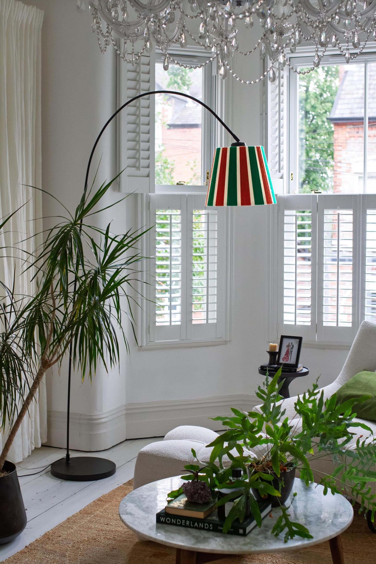 A tall black floor lamp with A Slice Shades handwoven yarn striped lampshade in Rust, Ecru & Jade Green standing in a modern living room next to a white sofa and potted plants.